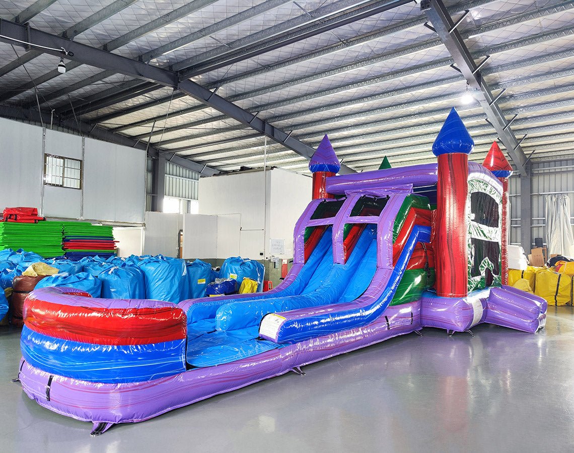 Wide-angle view of Multi Color Marble Splash & Save Dual Lane Inflatable Combo in a spacious warehouse. The inflatable displays vibrant purple, blue, and red colors with castle towers, dual slides, and a large splash pool. Various inflatable products, storage bags, and warehouse equipment are visible, demonstrating its size and commercial setting.