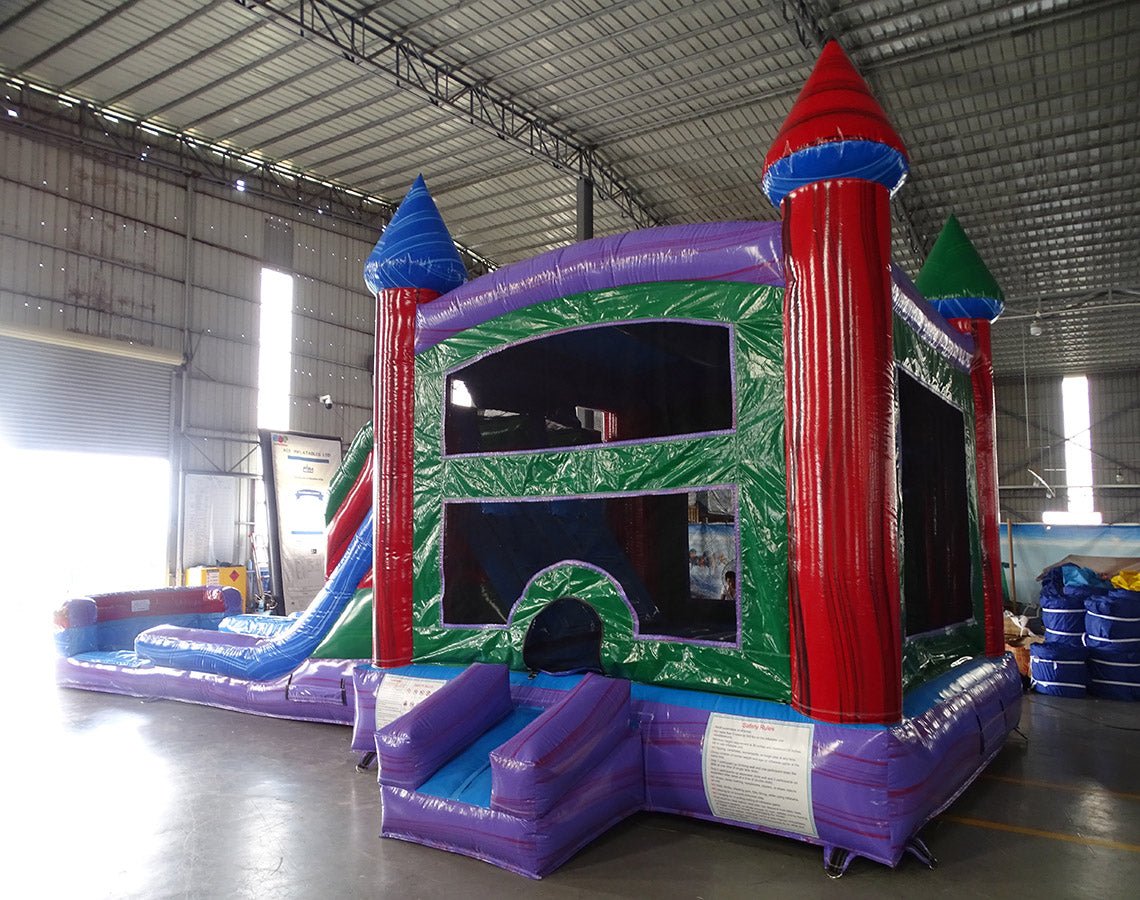 Cross Rear view of Multi Color Marble Splash & Save Dual Lane Inflatable Combo set up in a large indoor warehouse. The inflatable features colorful castle towers, dual slides, and a bounce house area with mesh windows. A green panel with white designs is visible on one side. Metal roofing and various inflatable products are visible in the background.