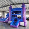 Side view of Mystic Castle Commercial Econo Inflatable Combo (Wet/Dry) set up in a large indoor warehouse. Features purple turrets, blue walls, yellow accents, slide, and splash pool. Metal roofing and other inflatable products visible in the background, demonstrating its size and indoor setup capabilities.