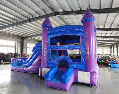 Side view of Mystic Castle Commercial Econo Inflatable Combo (Wet/Dry) set up in a large indoor warehouse. Features purple turrets, blue walls, yellow accents, slide, and splash pool. Metal roofing and other inflatable products visible in the background, demonstrating its size and indoor setup capabilities.