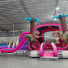 side view of Pink Dino Dive Dual Lane Commercial Inflatable Combo featuring a prehistoric theme. The structure has dual slides in pink and blue, emerging from a purple and pink base with inflatable palm trees. Two large green dinosaur figures flank the entrance. Set up in a large indoor warehouse with metal roofing and bright lighting visible.