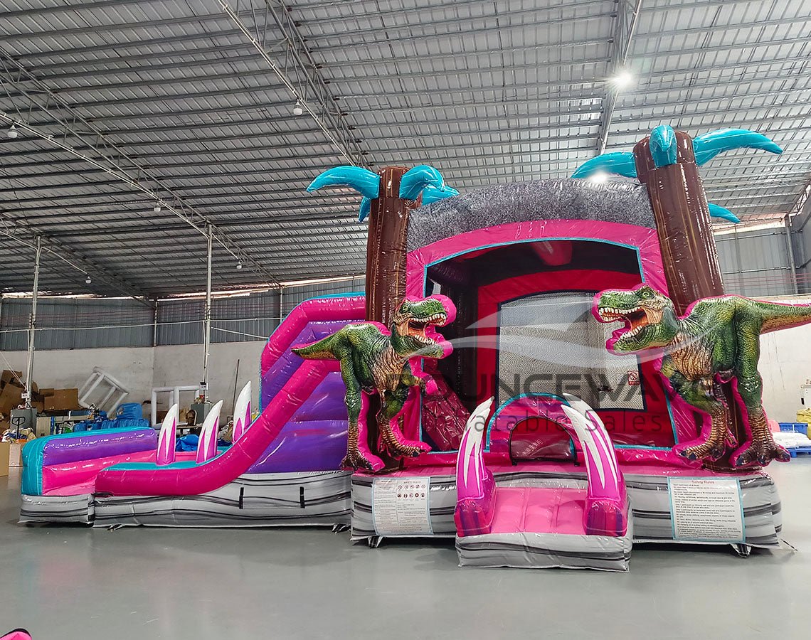 side view of Pink Dino Dive Dual Lane Commercial Inflatable Combo featuring a prehistoric theme. The structure has dual slides in pink and blue, emerging from a purple and pink base with inflatable palm trees. Two large green dinosaur figures flank the entrance. Set up in a large indoor warehouse with metal roofing and bright lighting visible.