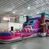 Wide-angle view of Pink Dino Dive Dual Lane Commercial Inflatable Combo in a spacious warehouse. The inflatable displays a vibrant pink and purple design with dual slides, inflatable palm trees, and green dinosaur figures. A large splash pool area is visible at the base. Various other inflatable products and warehouse equipment can be seen in the background.