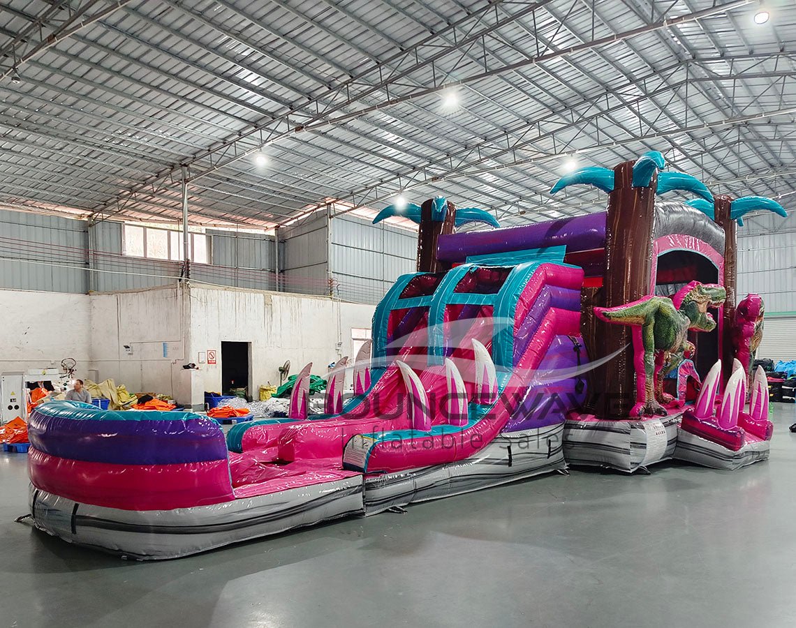 Wide-angle view of Pink Dino Dive Dual Lane Commercial Inflatable Combo in a spacious warehouse. The inflatable displays a vibrant pink and purple design with dual slides, inflatable palm trees, and green dinosaur figures. A large splash pool area is visible at the base. Various other inflatable products and warehouse equipment can be seen in the background.