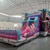 Three-quarter view of Pink Dino Dive Dual Lane Commercial Inflatable Combo set up in a warehouse. The structure features a pink and purple base with inflatable palm trees, dual slides, and large green dinosaur figures at the entrance. A colorful splash pool and various inflatable obstacles are visible. The image showcases the combo's size and detailed prehistoric theme in a commercial setting.