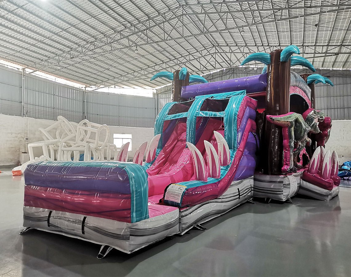 Three-quarter view of Pink Dino Dive Dual Lane Commercial Inflatable Combo set up in a warehouse. The structure features a pink and purple base with inflatable palm trees, dual slides, and large green dinosaur figures at the entrance. A colorful splash pool and various inflatable obstacles are visible. The image showcases the combo's size and detailed prehistoric theme in a commercial setting.