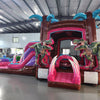 Large inflatable dinosaur-themed combo featuring a bounce house with palm tree decorations, attached slide, and splash pool. The structure has vibrant pink, purple, and blue colors with realistic T-Rex dinosaur figures guarding the entrance. Set up in an indoor warehouse with metal roofing visible.