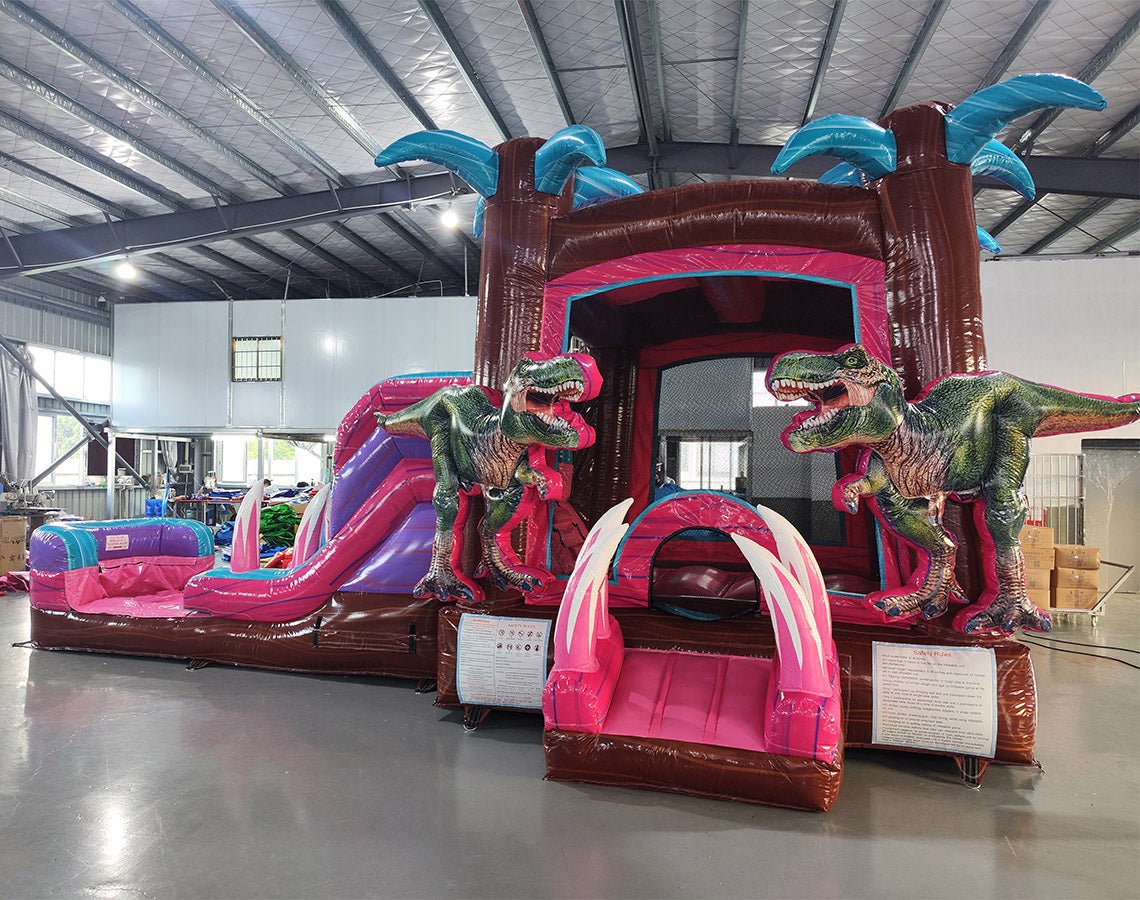 Large inflatable dinosaur-themed combo featuring a bounce house with palm tree decorations, attached slide, and splash pool. The structure has vibrant pink, purple, and blue colors with realistic T-Rex dinosaur figures guarding the entrance. Set up in an indoor warehouse with metal roofing visible.