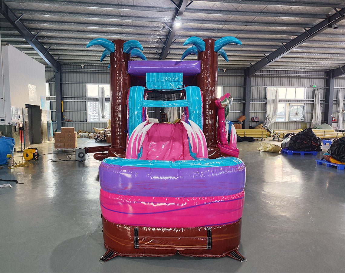 Front view of Pink Dino Econo Combo, showcasing the bounce house entrance with palm tree decorations, dinosaur figures, pink slide, and purple splash pool area. Set up in an indoor facility with other inflatable products and equipment visible in the background.