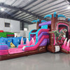 Wide-angle view of Pink Dino Econo Combo featuring a colorful bounce house with palm tree decorations, dinosaur figures, curved slide, and spacious splash pool. The inflatable is set up in an indoor warehouse with metal roofing, bright lighting, and other equipment visible in the background.