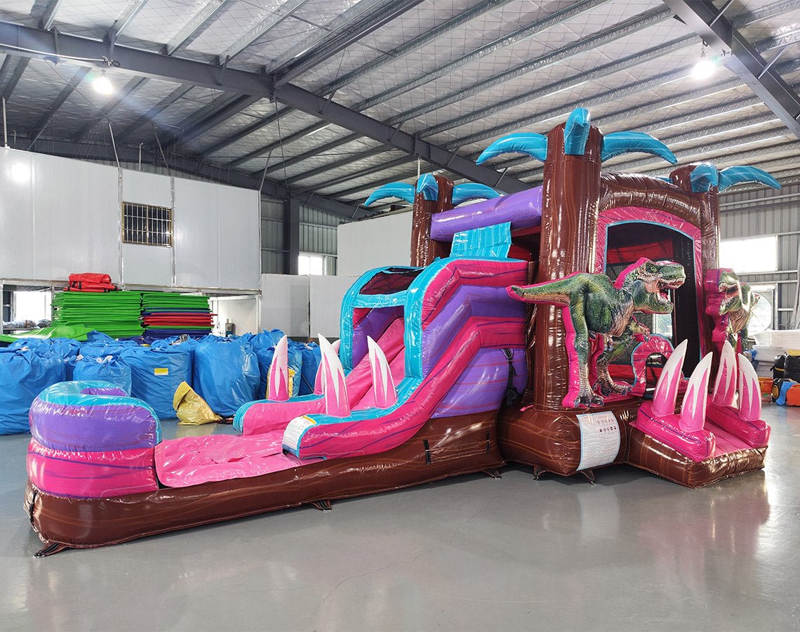 Wide-angle view of Pink Dino Econo Combo featuring a colorful bounce house with palm tree decorations, dinosaur figures, curved slide, and spacious splash pool. The inflatable is set up in an indoor warehouse with metal roofing, bright lighting, and other equipment visible in the background.