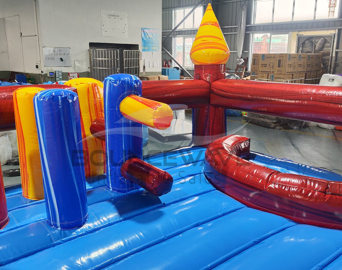 Close-up view of Primary Marble Toddler Bounce House interior, showing colorful inflatable obstacles including yellow and blue cylindrical pillars, a red arched tunnel, and a blue curved slide. The bounce floor is visible with a blue base. Set in a warehouse environment with metal roofing and windows in the background.