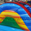 Side view of Primary Marble Toddler Bounce House featuring a vibrant design with a central yellow dome, blue base, and multicolored corner towers. The structure includes mesh windows for visibility and safety. Set up in a large indoor warehouse with metal roofing, windows, and other inflatable products visible in the background, showcasing its commercial setting.