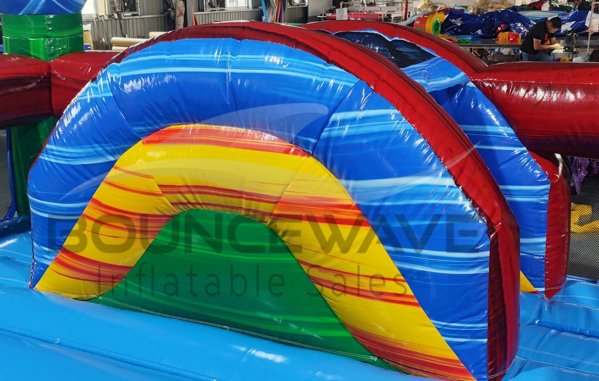 Side view of Primary Marble Toddler Bounce House featuring a vibrant design with a central yellow dome, blue base, and multicolored corner towers. The structure includes mesh windows for visibility and safety. Set up in a large indoor warehouse with metal roofing, windows, and other inflatable products visible in the background, showcasing its commercial setting.