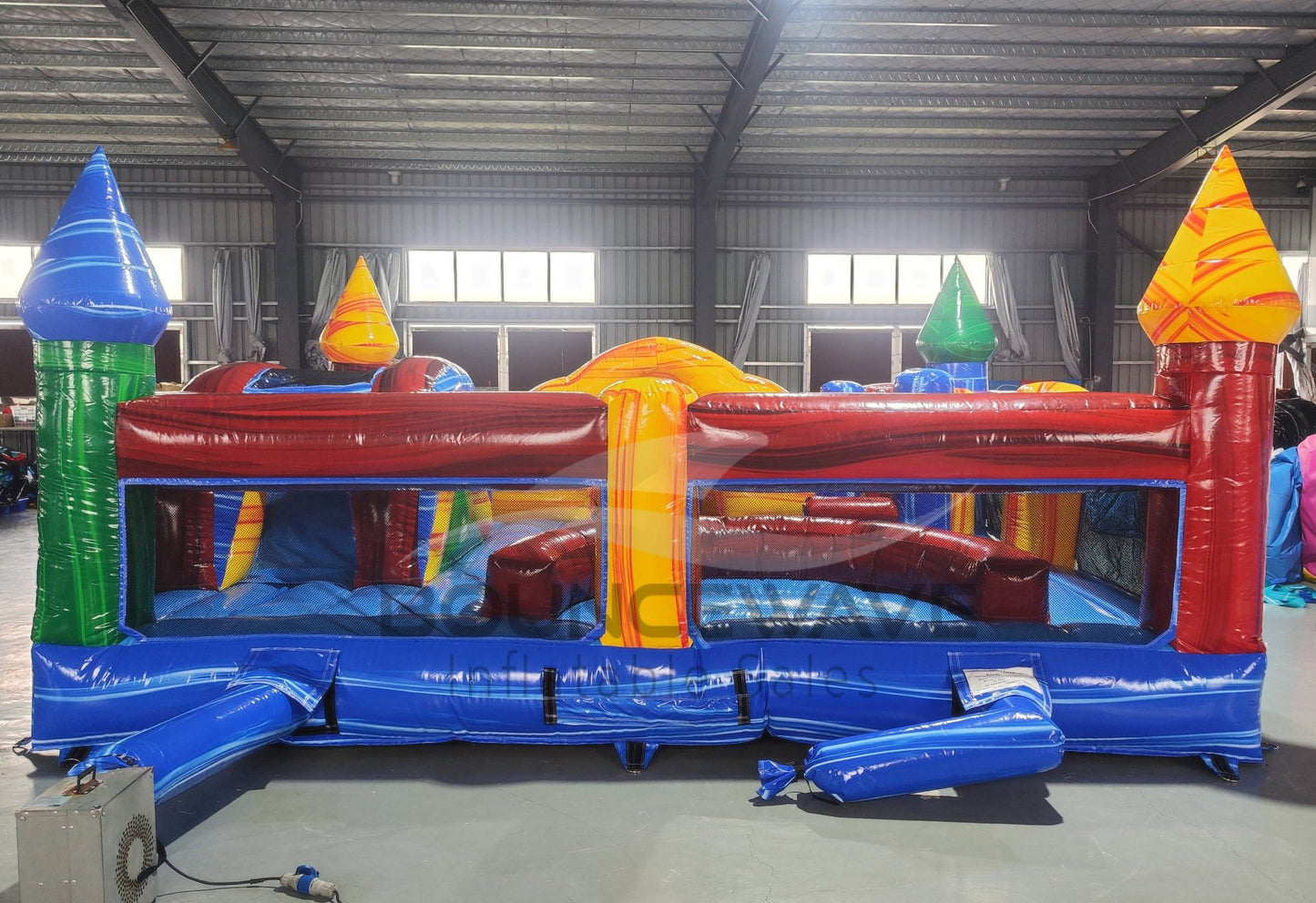 Rear view of Primary Marble Toddler Bounce House showing the blue base, red and maroon walls, and yellow dome top with orange swirl patterns. Four corner towers in various colors visible. Set in a warehouse with metal ceiling, windows, and other inflatable products in the background.