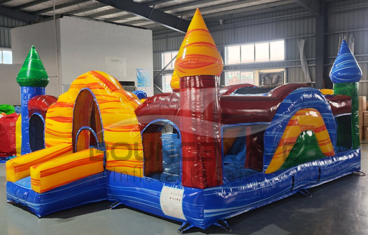 Colorful inflatable Primary Marble Toddler Bounce House featuring a central yellow dome with orange swirl patterns. Four corner towers in blue, green, red, and yellow with conical tops. Blue base with mesh windows and a small entrance slide. Set up in an indoor warehouse with metal roofing and windows visible.