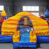 Wide-angle front view of Primary Marble Toddler Bounce House in a warehouse setting. Showcases the yellow dome with orange swirls, colorful corner towers, blue base, and entrance slide. Metal roofing, windows, and other inflatable products visible in the background, providing context for size and commercial use.