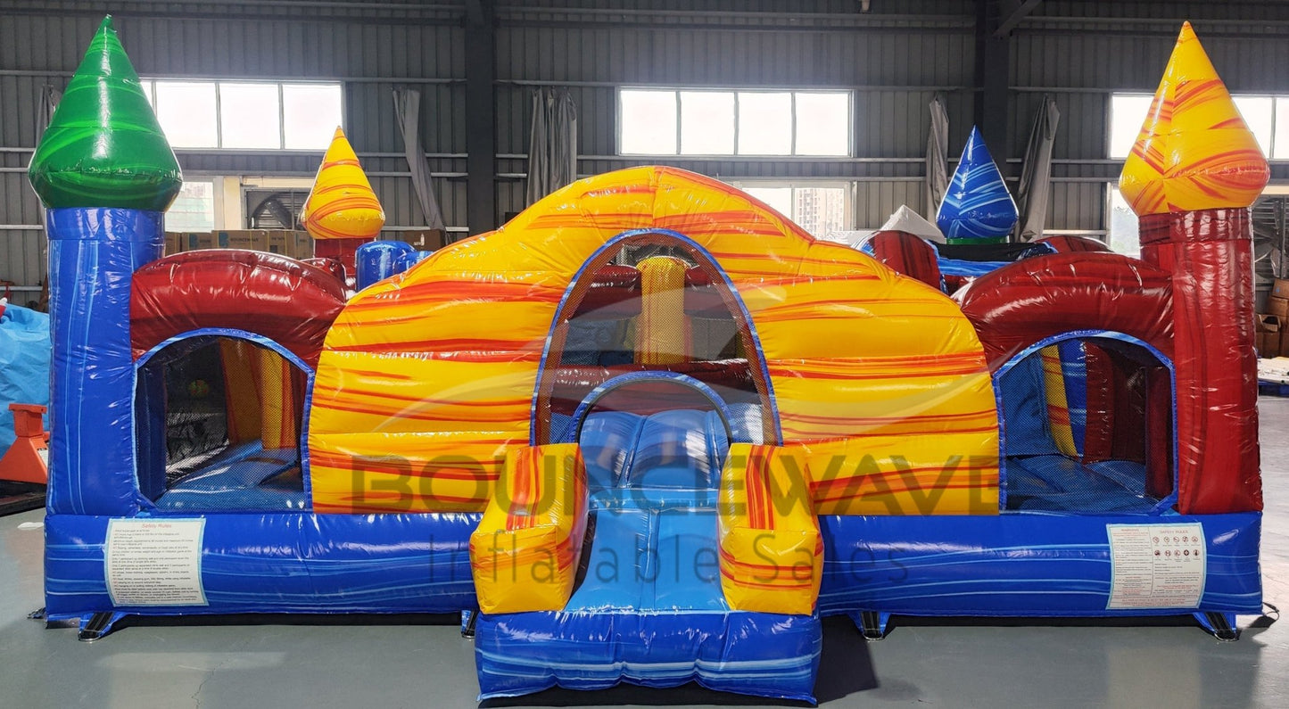 Wide-angle front view of Primary Marble Toddler Bounce House in a warehouse setting. Showcases the yellow dome with orange swirls, colorful corner towers, blue base, and entrance slide. Metal roofing, windows, and other inflatable products visible in the background, providing context for size and commercial use.