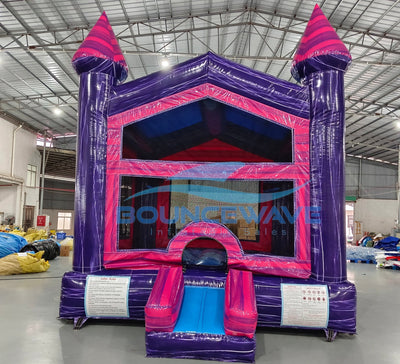 Wide-angle front view of Purple Palace Commercial Grade Bounce House in an indoor facility. The purple and pink inflatable features two tall towers, mesh windows, and a small front slide. Safety instructions are visible on the base. The warehouse setting shows metal roofing, fluorescent lighting, and various inflatable products and equipment in the background, demonstrating its commercial use context.