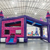 Wide-angle front view of Purple Palace Commercial Grade Canopy Bounce House in a spacious warehouse. The purple and pink inflatable features four tall towers with conical tops, a canopy roof, and a small front slide. The entrance shows a pink interior. Various inflatable products, equipment, and warehouse features are visible in the background, demonstrating its commercial use context.