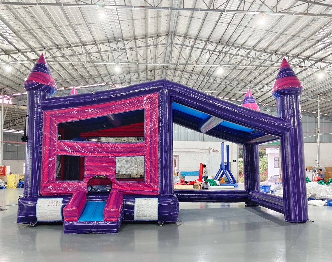 Wide-angle front view of Purple Palace Commercial Grade Canopy Bounce House in a spacious warehouse. The purple and pink inflatable features four tall towers with conical tops, a canopy roof, and a small front slide. The entrance shows a pink interior. Various inflatable products, equipment, and warehouse features are visible in the background, demonstrating its commercial use context.