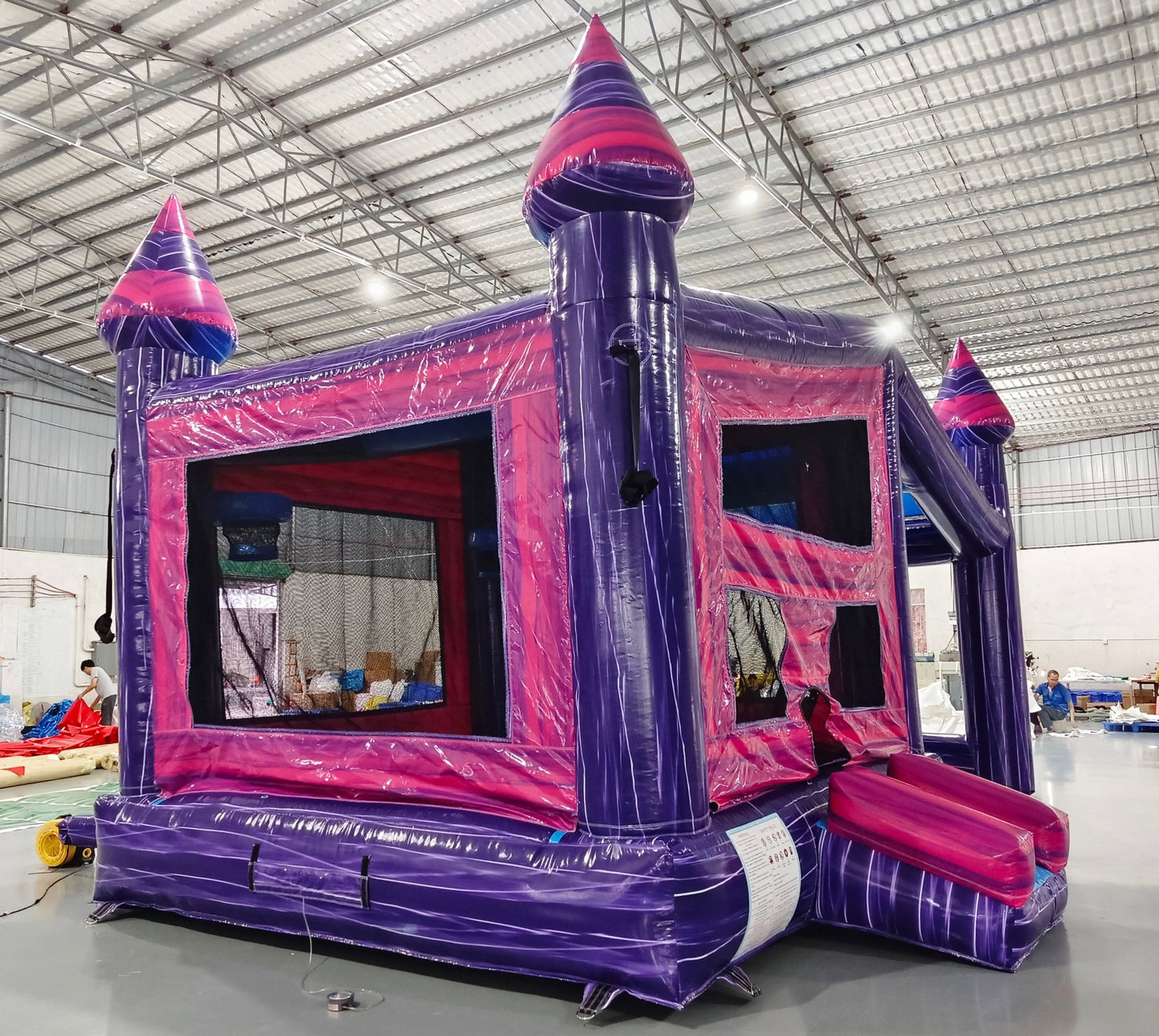 Three-quarter view of Purple Palace Commercial Grade Canopy Bounce House in an indoor facility. The purple inflatable structure has pink accents, four tall towers with conical tops, and a canopy roof. Mesh windows and a small slide are visible. The warehouse setting shows metal roofing, fluorescent lighting, and various inflatable products and equipment in the background, showcasing its size and commercial application.