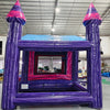 Rear view of Purple Palace Commercial Grade Canopy Bounce House in a large indoor warehouse. The inflatable structure features a vibrant purple exterior with pink accents. It has four tall purple towers with pink conical tops, a pink entrance with mesh windows, and a small slide at the front. The canopy extends between the towers, creating a covered play area. Safety instructions are visible on the base. 