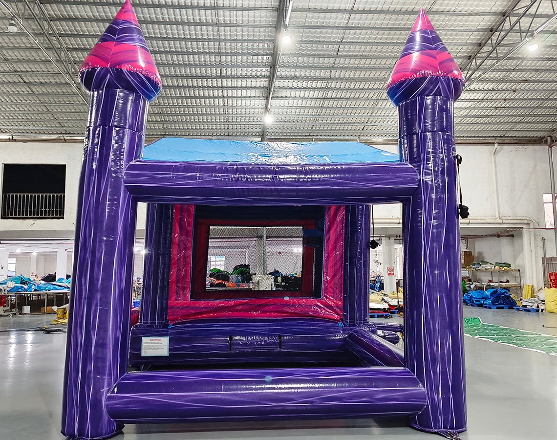 Rear view of Purple Palace Commercial Grade Canopy Bounce House in a large indoor warehouse. The inflatable structure features a vibrant purple exterior with pink accents. It has four tall purple towers with pink conical tops, a pink entrance with mesh windows, and a small slide at the front. The canopy extends between the towers, creating a covered play area. Safety instructions are visible on the base. 