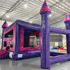 Side angle view of Purple Palace Commercial Grade Canopy Bounce House set up in a large indoor warehouse. The inflatable features purple walls, pink interior, and three tall towers with pink conical tops. A canopy extends between the towers, creating a covered play area. The warehouse setting shows metal roofing, fluorescent lighting, and other inflatable products in the background.