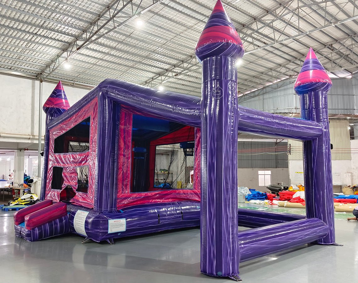 Side angle view of Purple Palace Commercial Grade Canopy Bounce House set up in a large indoor warehouse. The inflatable features purple walls, pink interior, and three tall towers with pink conical tops. A canopy extends between the towers, creating a covered play area. The warehouse setting shows metal roofing, fluorescent lighting, and other inflatable products in the background.