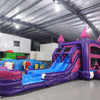 Front view of Purple Plunge Splash & Save Dual Lane Inflatable Combo featuring a castle-themed design with purple walls and pink turrets. The structure includes dual blue slides, a bounce house area with mesh windows, and a circular splash pool. Set up in a large indoor warehouse with metal roofing and bright lighting visible.