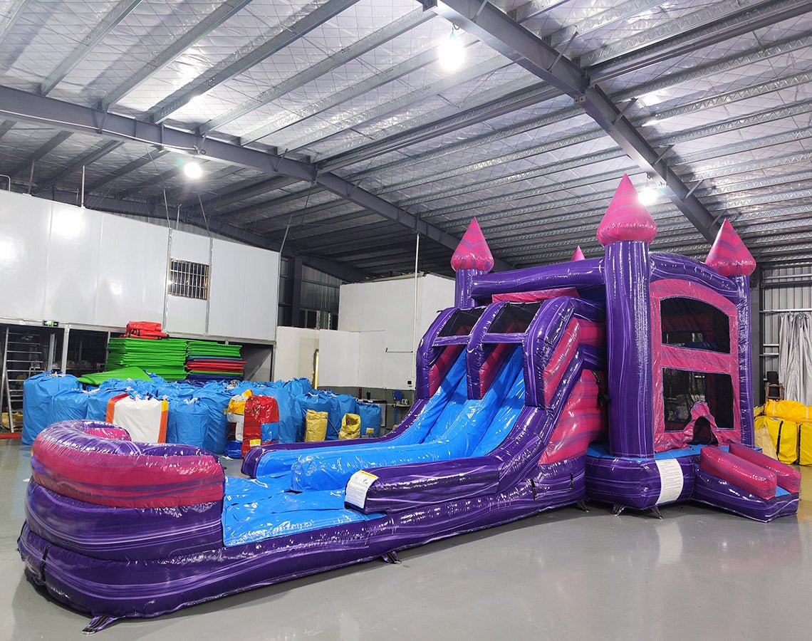 Front view of Purple Plunge Splash & Save Dual Lane Inflatable Combo featuring a castle-themed design with purple walls and pink turrets. The structure includes dual blue slides, a bounce house area with mesh windows, and a circular splash pool. Set up in a large indoor warehouse with metal roofing and bright lighting visible.