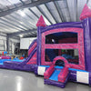 Side view of Purple Plunge Splash & Save Dual Lane Inflatable Combo showing dual blue slides emerging from a purple and pink castle structure. The inflatable features pink turrets with conical tops, a bounce house area, and a large circular splash pool. Various other inflatable products and warehouse equipment are visible in the background.