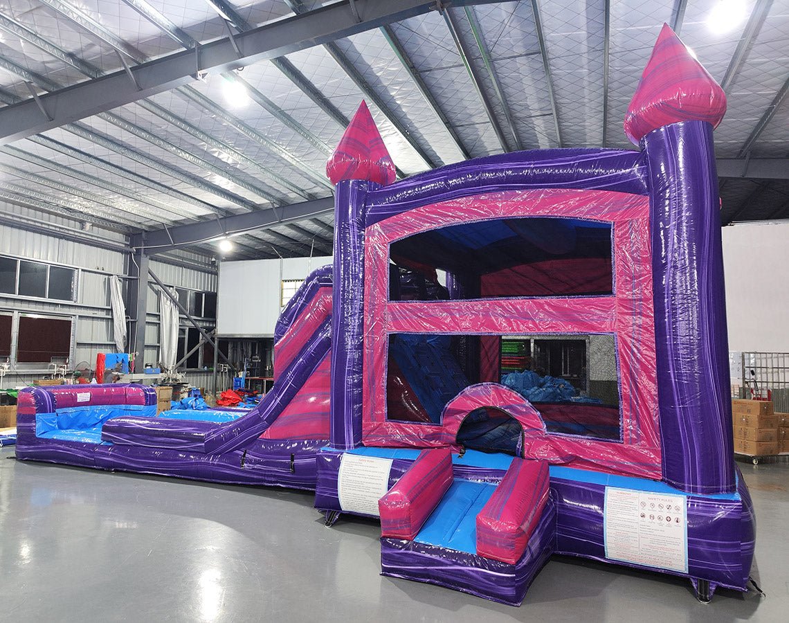 Side view of Purple Plunge Splash & Save Dual Lane Inflatable Combo showing dual blue slides emerging from a purple and pink castle structure. The inflatable features pink turrets with conical tops, a bounce house area, and a large circular splash pool. Various other inflatable products and warehouse equipment are visible in the background.