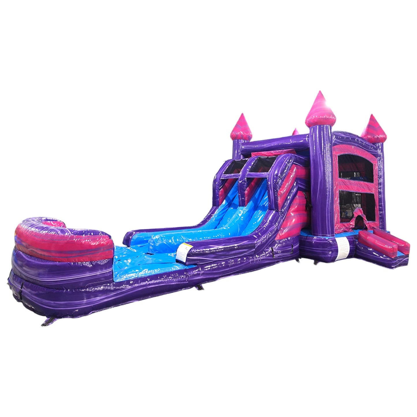 Side view of Purple Plunge Splash & Save Dual Lane Inflatable Combo isolated on a white background. The inflatable features a purple castle structure with pink turrets and conical tops, dual blue slides, a bounce house area, and a circular splash pool. The design showcases its castle theme for commercial wet/dry use.