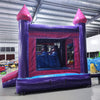 Rear Wide-angle view of Purple Plunge Splash & Save Dual Lane Inflatable Combo (Wet/Dry) set up in a large indoor warehouse. The inflatable features a castle-like structure with purple walls, pink conical turrets, and dual blue slides. It includes a bounce house area with mesh windows and a circular splash pool. Various colorful inflatable products and equipment are visible in the background, showcasing the warehouse setting with metal roofing and bright lighting.