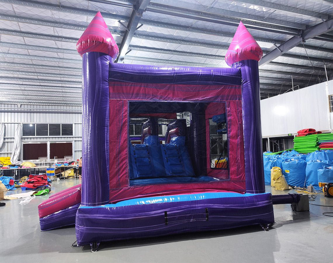 Rear Wide-angle view of Purple Plunge Splash & Save Dual Lane Inflatable Combo (Wet/Dry) set up in a large indoor warehouse. The inflatable features a castle-like structure with purple walls, pink conical turrets, and dual blue slides. It includes a bounce house area with mesh windows and a circular splash pool. Various colorful inflatable products and equipment are visible in the background, showcasing the warehouse setting with metal roofing and bright lighting.