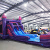 Wide-angle view of Purple Plunge Splash & Save Dual Lane Inflatable Combo in a spacious warehouse. The inflatable displays a purple castle design with pink accents, dual blue slides, and a large splash pool area. Other inflatable products, storage bags, and warehouse materials can be seen, demonstrating its size and commercial setting.