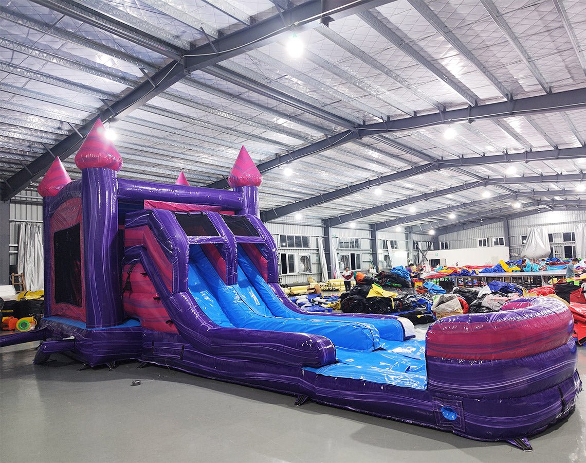 Wide-angle view of Purple Plunge Splash & Save Dual Lane Inflatable Combo in a spacious warehouse. The inflatable displays a purple castle design with pink accents, dual blue slides, and a large splash pool area. Other inflatable products, storage bags, and warehouse materials can be seen, demonstrating its size and commercial setting.