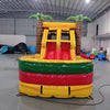 Front view of Rasta Splash Dual Lane Commercial Inflatable Combo set up in a warehouse. The inflatable displays dual slides in red and yellow, inflatable palm trees, and a circular splash pool area in vibrant colors of yellow, green, and red. Other inflatable products and warehouse materials are visible in the background, demonstrating its commercial setting.