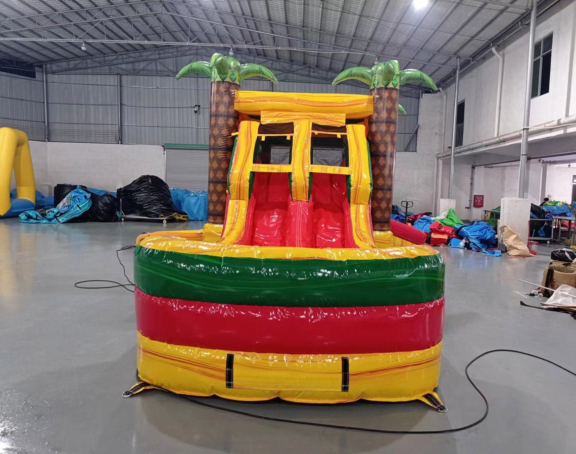 Front view of Rasta Splash Dual Lane Commercial Inflatable Combo set up in a warehouse. The inflatable displays dual slides in red and yellow, inflatable palm trees, and a circular splash pool area in vibrant colors of yellow, green, and red. Other inflatable products and warehouse materials are visible in the background, demonstrating its commercial setting.