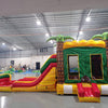 Side view of Rasta Splash Dual Lane Commercial Inflatable Combo showing dual slides in red and yellow emerging from a green structure with inflatable palm trees. The combo includes a bounce house area and a large splash pool. Various other inflatable products and warehouse equipment are visible in the background.