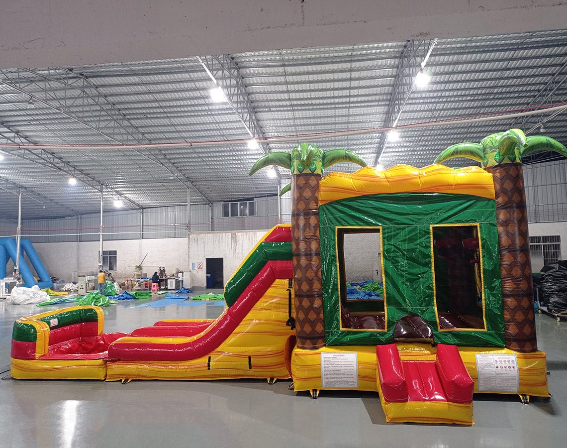 Side view of Rasta Splash Dual Lane Commercial Inflatable Combo showing dual slides in red and yellow emerging from a green structure with inflatable palm trees. The combo includes a bounce house area and a large splash pool. Various other inflatable products and warehouse equipment are visible in the background.