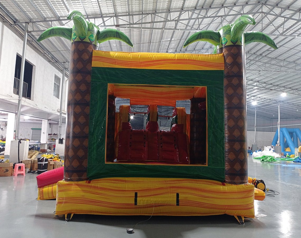 Rear view of Rasta Splash Dual Lane Commercial Inflatable Combo featuring a tropical theme with inflatable palm trees. The structure has dual slides in red and yellow, a green bounce house area with mesh windows, and a colorful base in yellow, green, and red. Set up in a large indoor warehouse with metal roofing and bright lighting visible.