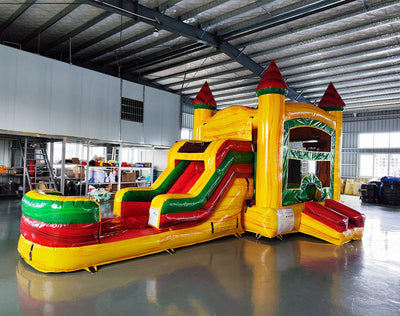 Wide-angle view of Rasta Splash Commercial Econo Inflatable Combo (Wet/Dry) in an indoor warehouse setting. The inflatable features a castle-themed bounce house with yellow walls, red turrets, green accents, a curved slide, and a splash pool area. Other inflatable products, shelving units, and warehouse equipment visible in the background, demonstrating its size and commercial setting.