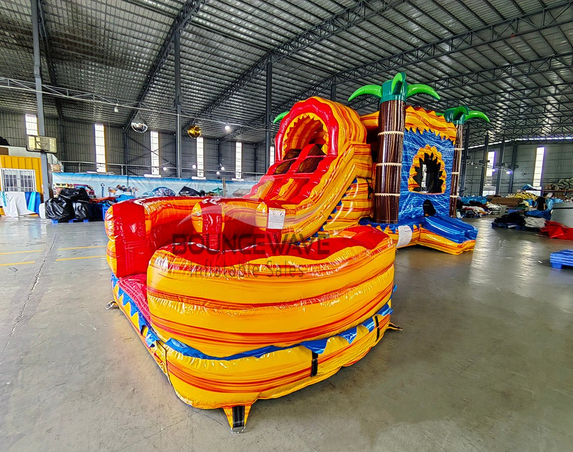 Front view of Rasta Curved Dual Lane Commercial Inflatable Combo (Wet/Dry) set up in a large indoor warehouse. Vibrant inflatable with dual red and orange curved slides, blue castle-like entrance, and palm tree decorations. Large splash pool visible. Metal roofing, disco balls, and other warehouse equipment provide context for its commercial use and size.