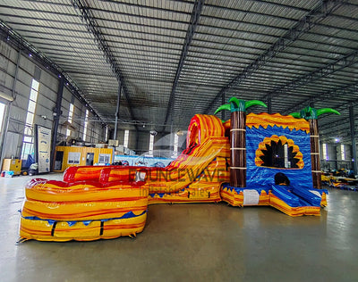 Wide-angle view of Rasta Curved Dual Lane Commercial Inflatable Combo (Wet/Dry) in a spacious warehouse setting. Showcases the full structure including dual curved slides, blue castle entrance, palm trees, and large splash pool. Other inflatable products, shelving units, and warehouse equipment visible, demonstrating its scale in a commercial environment.