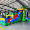 Wide-angle view of Reggae Rush Dual Lane Commercial Inflatable Combo in a spacious warehouse. The inflatable displays green palm trees, dual blue slides, a yellow and green bounce house structure, and a large colorful splash pool area. Various other inflatable products and warehouse equipment are visible, showcasing its size and commercial setting.