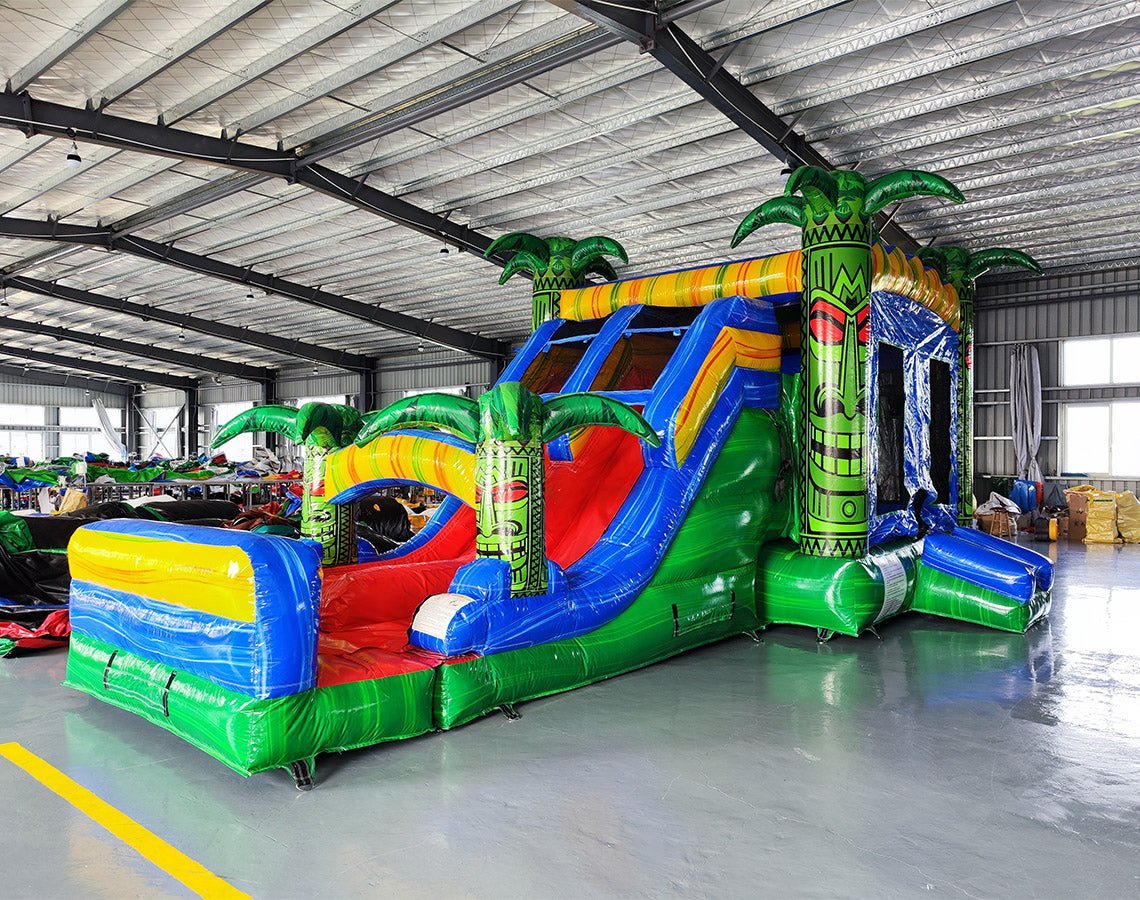 Wide-angle view of Reggae Rush Dual Lane Commercial Inflatable Combo in a spacious warehouse. The inflatable displays green palm trees, dual blue slides, a yellow and green bounce house structure, and a large colorful splash pool area. Various other inflatable products and warehouse equipment are visible, showcasing its size and commercial setting.