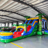 Three-quarter view of Reggae Rush Dual Lane Commercial Inflatable Combo in a warehouse environment. The inflatable features green palm trees, dual blue slides, a yellow and green bounce house area, and a vibrant splash pool. Other inflatable products and warehouse materials in the background demonstrate its commercial application.